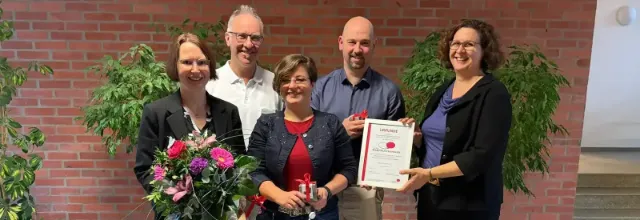 Eine Gruppe von fünf lächelnden Menschen steht in einem Raum auf einem karierten Boden. Einer hält einen Blumenstrauss, ein anderer eine Urkunde. Im Hintergrund sind Grünpflanzen und eine Backsteinwand zu sehen.