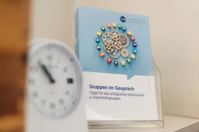 Nahaufnahme einer kleinen Uhr im Vordergrund, unscharf, vor einer blauen Broschüre von Selbsthilfe BE.