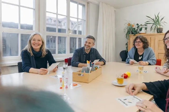 Vier Personen sitzen an einem hellen Holztisch in einem freundlichen Besprechungsraum und arbeiten gemeinsam an einer Selbsthilfegruppen-Arbeit Stiften und Karten