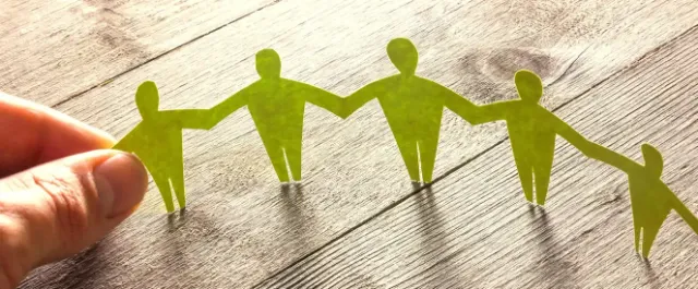 Hand hält eine Papierkette aus grünen Figuren, die sich an den Händen halten, auf einem Holztisch liegend.