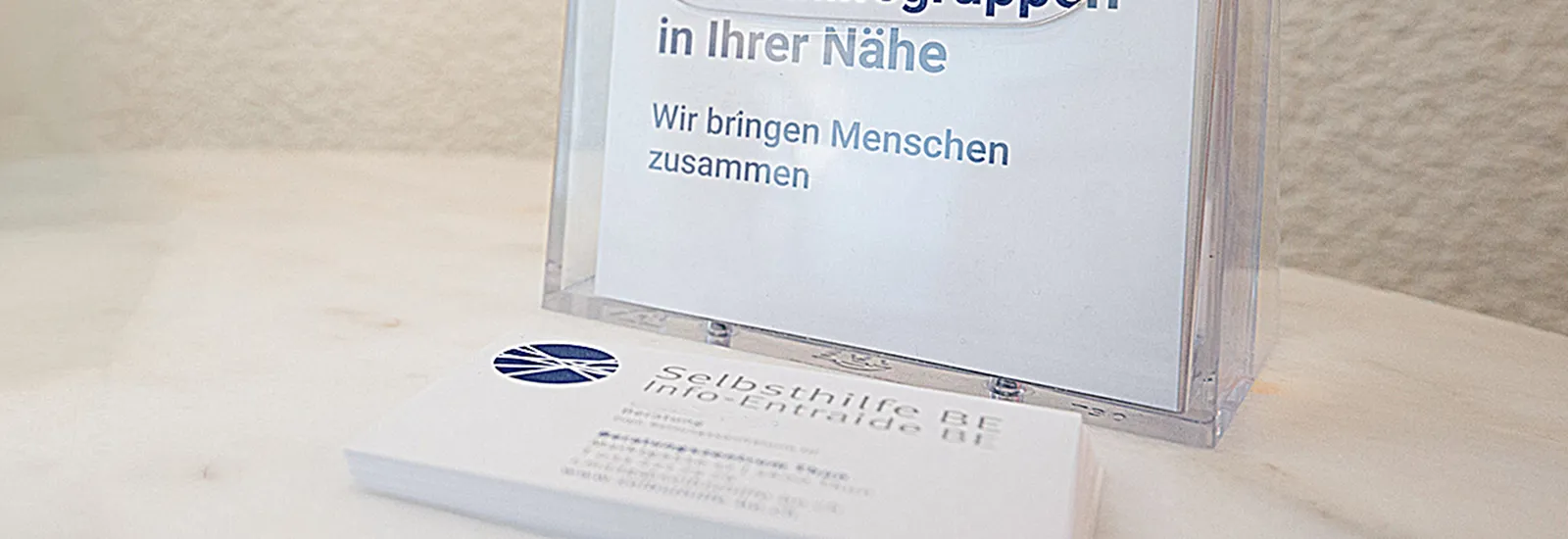 Ein Stapel Visitenkarten der Organisation „Selbsthilfe BE“ liegt auf einem hellen Holztisch. Dahinter steht ein durchsichtiger Acryl-Ständer mit einem Einleger, auf dem in grosser Schrift „in Ihrer Nähe“ und darunter „Wir bringen Menschen zusammen“ steht.
