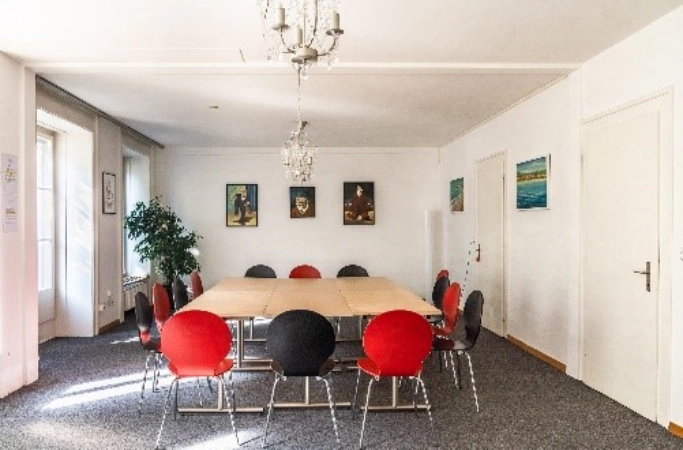 Gruppenraum mit rechteckigen Tischen in U-Form, rot-schwarzen Stühlen, Wandbildern, Teppichboden und Kronleuchtern.