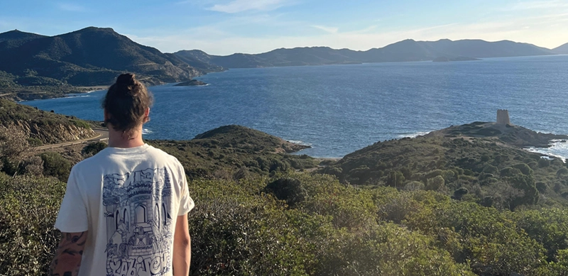 Person mit Dutt und T-Shirt mit großem Motiv steht mit dem Rücken zur Kamera und blickt auf eine Küstenlandschaft mit Hügeln, Meer und einem Turm auf einer Landzunge