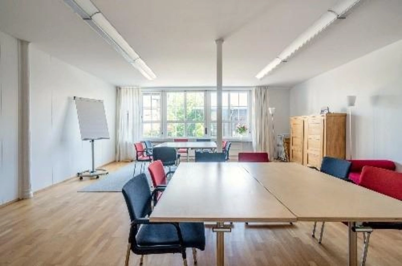 Moderner Seminarraum mit Holzboden, grossen Fenstern, rechteckigem Tisch, verschiedenfarbigen Stühlen und Flipchart.