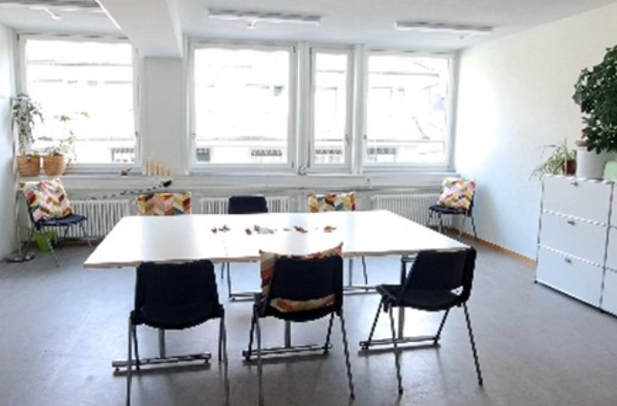 Heller Gruppenraum mit grossem rechteckigem Tisch, sechs schwarzen Stühlen, bunten Kissen auf Fensterbänken und Zimmerpflanzen.