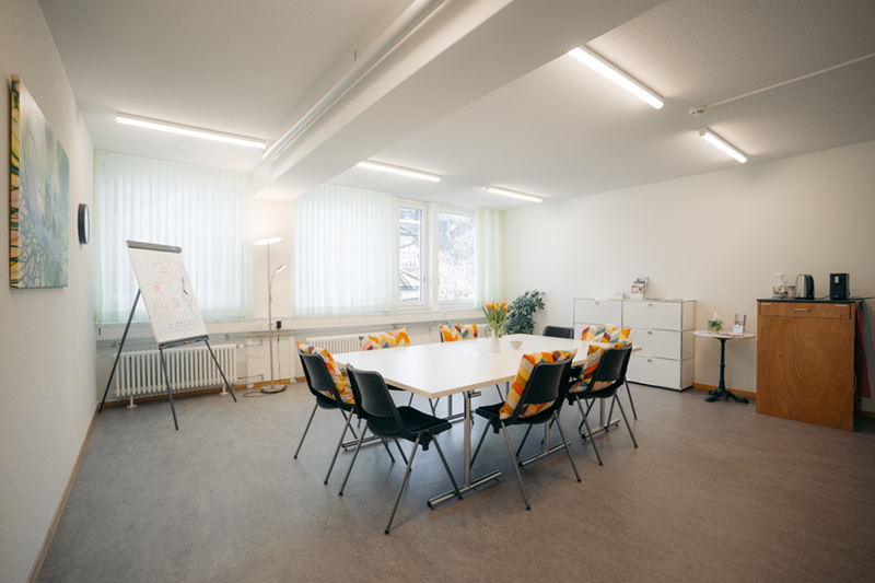 Heller Gruppenraum mit grossem rechteckigem Tisch, sechs schwarzen Stühlen, bunten Kissen auf Fensterbänken und Zimmerpflanzen.