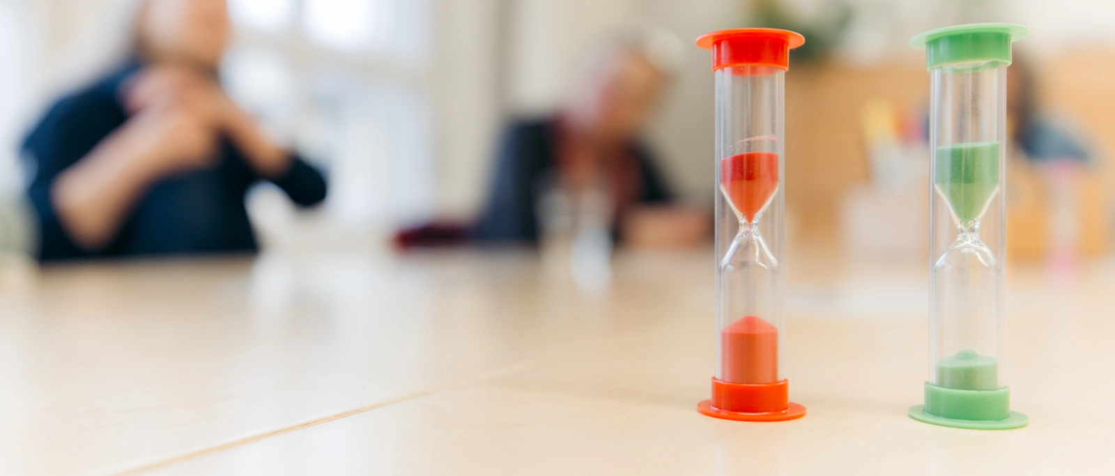 Zwei Sanduhren mit rotem und grünem Sand stehen auf einem Tisch vor unscharfen Personen im Hintergrund.