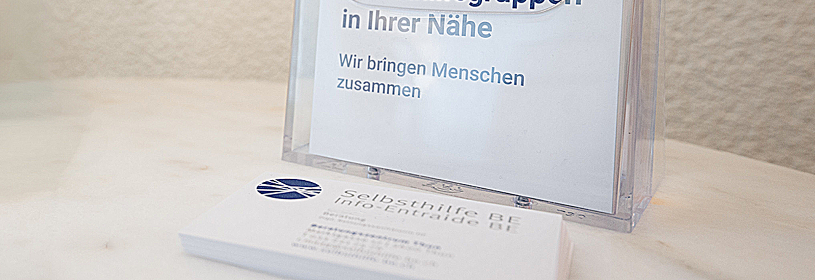 Ein Stapel Visitenkarten der Organisation „Selbsthilfe BE“ liegt auf einem hellen Holztisch. Dahinter steht ein durchsichtiger Acryl-Ständer mit einem Einleger, auf dem in grosser Schrift „in Ihrer Nähe“ und darunter „Wir bringen Menschen zusammen“ steht.